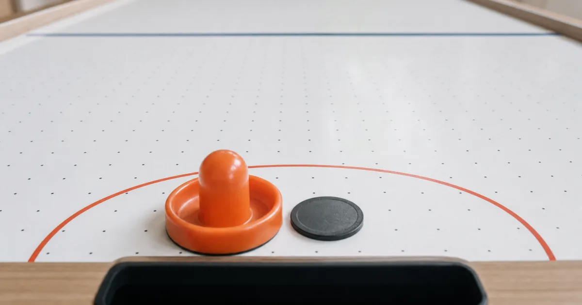 En orange kølle og en sort puck foran målet på et hvidt airhockeybord
